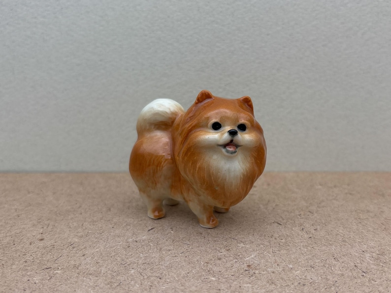 May include: A small, ceramic figurine of a brown and white Pomeranian dog. The dog is standing on all four paws and has a fluffy tail.