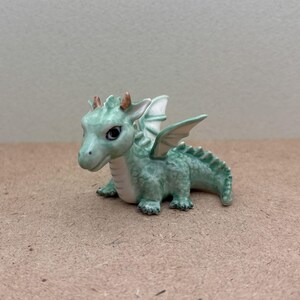 May include: A small green ceramic dragon figurine with gold accents and white wings. The dragon is sitting on its hind legs with its wings spread out.