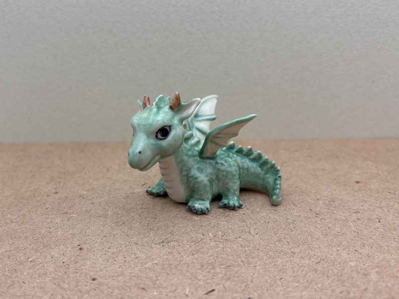 May include: A small green ceramic dragon figurine with gold accents and white wings. The dragon is sitting on its hind legs with its wings spread out.