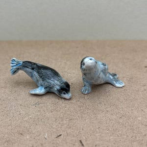 Może przedstawiać: Dwie małe ceramiczne figurki fok. Jedna foka jest szara i czarna z niebieską płetwą ogonową. Druga foka jest biała i szara z niebieską płetwą ogonową.