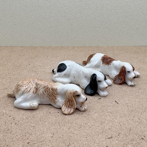 Peut inclure: Trois figurines de chiens en céramique en position de repos. Les chiens sont blancs avec des marques marron et noires. Les figurines ont des oreilles tombantes et sont couchées.
