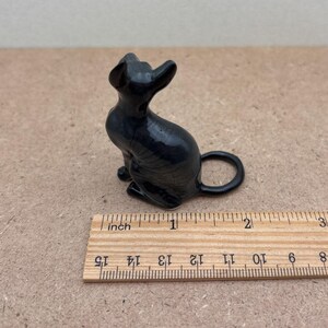 K&ouml;nnte beinhalten: Eine schwarze Keramikfigur eines sitzenden Katers mit dem Schwanz um die Beine gelegt. Die Figur ist etwa 2,5 cm hoch und steht auf einem Holzlineal.