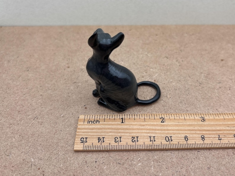 K&ouml;nnte beinhalten: Eine schwarze Keramikfigur eines sitzenden Katers mit dem Schwanz um die Beine gelegt. Die Figur ist etwa 2,5 cm hoch und steht auf einem Holzlineal.