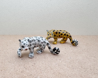 Puede incluir: Dos figuritas de cerámica de leopardos. Una es blanca con manchas negras y la otra es amarilla con manchas negras.