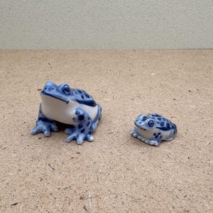 May include: Two ceramic frog ornaments. The larger frog is white with blue spots and accents. The smaller frog has a similar design. Both frogs are on a light brown surface.