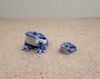Op de afbeelding: Twee keramische kikkerbeeldjes. De grotere kikker is wit met blauwe stippen en accenten. De kleinere kikker heeft een vergelijkbaar ontwerp. Beide kikkers staan op een lichtbruin oppervlak.