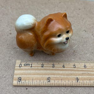 May include: A small ceramic figurine of a brown and white Pomeranian dog. The dog is sitting with its head tilted to the side and its tongue sticking out. The figurine is about 2 inches tall.
