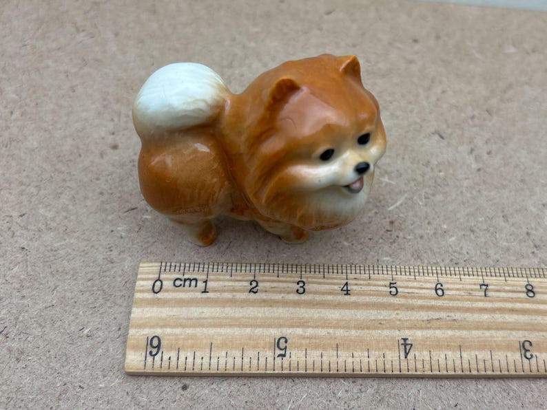 May include: A small ceramic figurine of a brown and white Pomeranian dog. The dog is sitting with its head tilted to the side and its tongue sticking out. The figurine is about 2 inches tall.