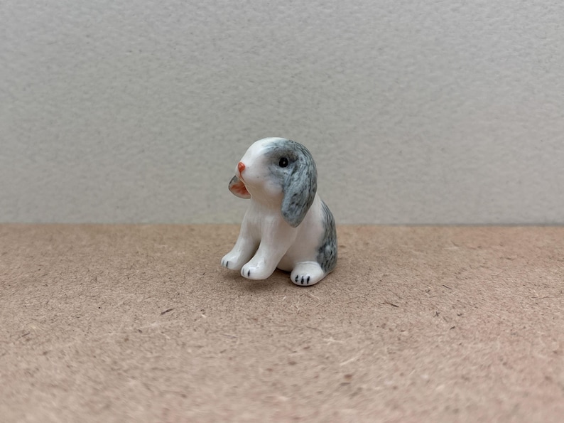 May include: A small ceramic figurine of a gray and white rabbit sitting with its ears perked up.