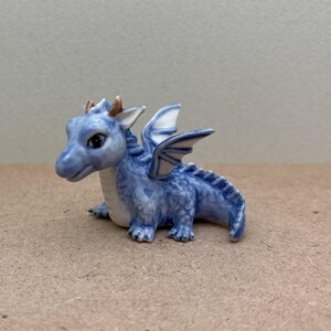 May include: A small blue ceramic dragon figurine with white wings and gold accents. The dragon is sitting on its hind legs with its wings spread out.
