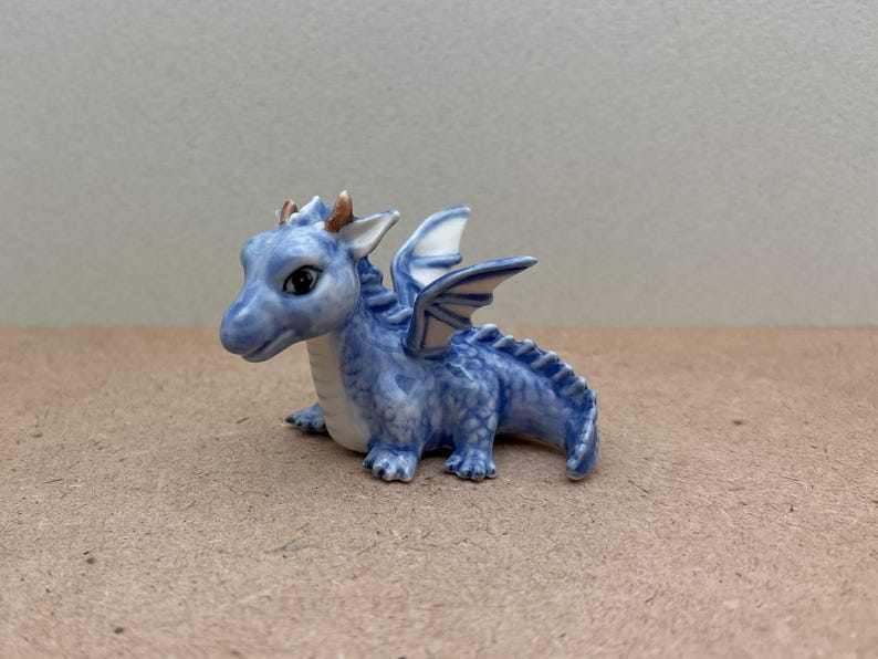 May include: A small blue ceramic dragon figurine with white wings and gold accents. The dragon is sitting on its hind legs with its wings spread out.