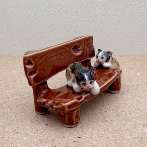May include: A brown ceramic bench with two small cat figurines. One cat is black and white, the other is calico. The bench has a rustic design with carved details and is a decorative item.