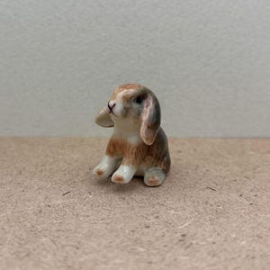 May include: A small ceramic figurine of a brown and white rabbit sitting with its ears flopped down. The rabbit has a white face and brown spots on its body.