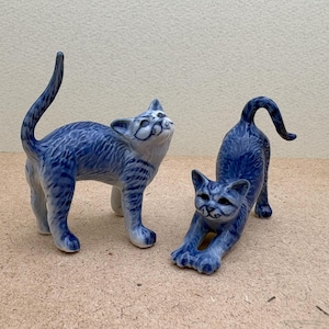 May include: Two blue and white ceramic cat figurines. One cat is standing on all fours with its head tilted up. The other cat is stretching with its front paws extended forward.