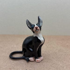 Puede incluir: Una figurita de cer&aacute;mica de un gato, pintada de blanco y negro, sentada con la cabeza inclinada hacia arriba. El gato tiene ojos azules y patas rosas.