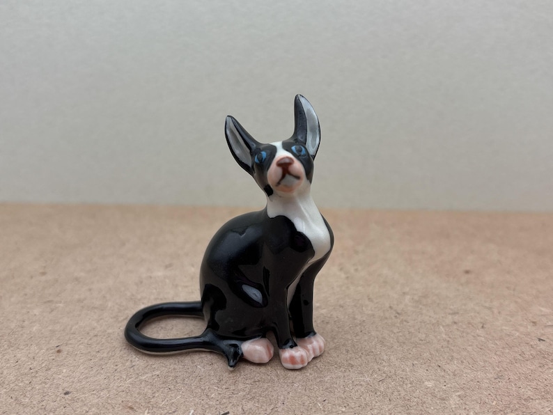 Puede incluir: Una figurita de cer&aacute;mica de un gato, pintada de blanco y negro, sentada con la cabeza inclinada hacia arriba. El gato tiene ojos azules y patas rosas.
