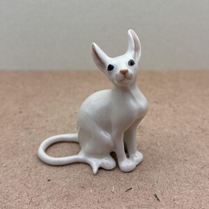 K&ouml;nnte beinhalten: Wei&szlig;e Porzellanfigur eines Katers mit blauen Augen. Der Kater sitzt mit seinem Schwanz um seinen K&ouml;rper gewickelt.