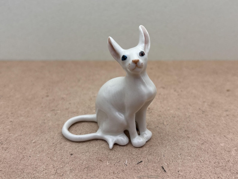 K&ouml;nnte beinhalten: Wei&szlig;e Porzellanfigur eines Katers mit blauen Augen. Der Kater sitzt mit seinem Schwanz um seinen K&ouml;rper gewickelt.