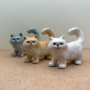 May include: Three ceramic cat figurines, one blue, one orange, and one white. The cats are all in a sitting position with their tails curled up.