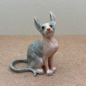 Puede incluir: Una peque&ntilde;a figurita de cer&aacute;mica de un gato sin pelo gris y rosa sentado con la cabeza levantada. El gato tiene ojos verdes y la nariz rosa.