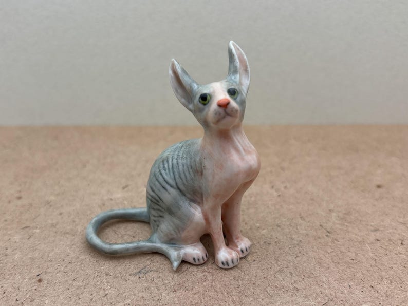 Puede incluir: Una peque&ntilde;a figurita de cer&aacute;mica de un gato sin pelo gris y rosa sentado con la cabeza levantada. El gato tiene ojos verdes y la nariz rosa.