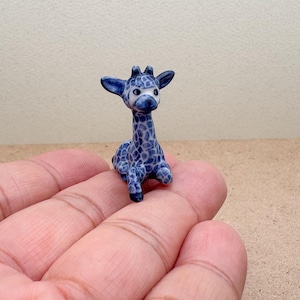 Puede incluir: Una pequeña figura de jirafa de cerámica en posición sentada. La jirafa tiene un patrón de manchas azules y blancas, con una cara blanca y orejas azul oscuro. La figura está en la mano de una persona.