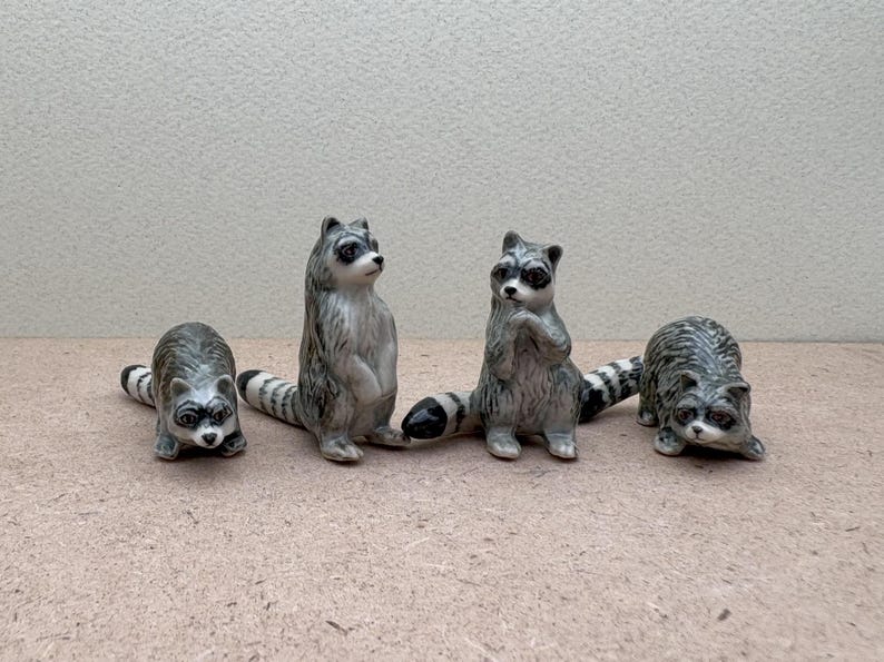 Pu&ograve; includere: Quattro statuine di procione in ceramica con code a strisce bianche e nere. I procioni sono in piedi su una superficie marrone chiaro.