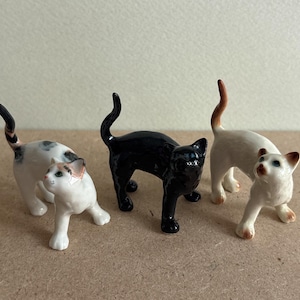 May include: Three ceramic cat figurines. One is black, one is white with black spots, and one is white with brown spots.