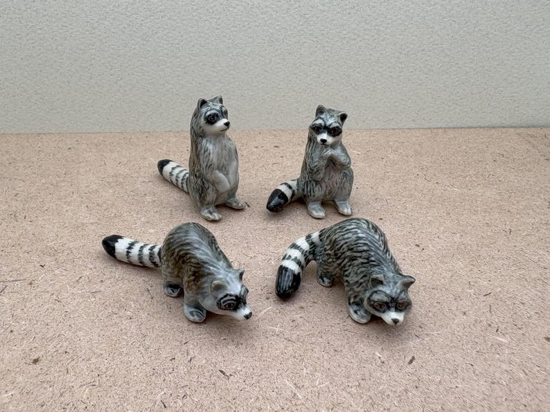 Pu&ograve; includere: Quattro statuine di procione in ceramica. I procioni sono grigi e neri con segni bianchi. Sono in piedi su una superficie marrone chiaro.