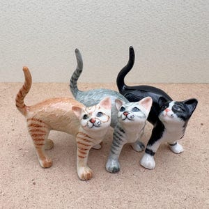 K&ouml;nnte beinhalten: Vier Keramik-Katzenfiguren in verschiedenen Farben: orange getigert, grau getigert, schwarz und schwarz-wei&szlig;. Jede Katze steht mit erhobenem Kopf. Die Figuren sind detailliert mit bemalten Merkmalen und Markierungen.
