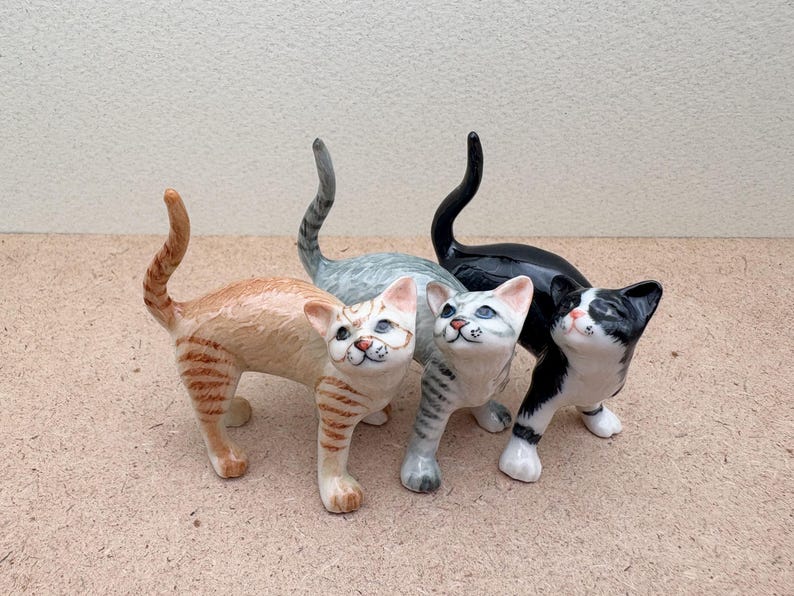 K&ouml;nnte beinhalten: Vier Keramik-Katzenfiguren in verschiedenen Farben: orange getigert, grau getigert, schwarz und schwarz-wei&szlig;. Jede Katze steht mit erhobenem Kopf. Die Figuren sind detailliert mit bemalten Merkmalen und Markierungen.