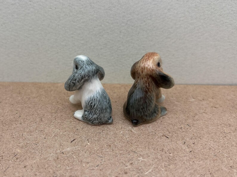 May include: Two small ceramic rabbit figurines. The rabbit on the left is white with gray markings. The rabbit on the right is brown with gray markings.
