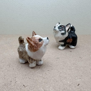 May include: Two ceramic corgi figurines. One is brown, white, and tan, the other is black, white, and brown. Both are standing and looking upwards. The figurines have detailed fur textures and are on a neutral surface.
