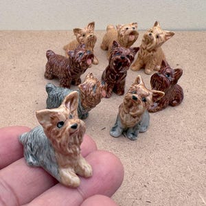 Puede incluir: Una colecci&oacute;n de nueve figuritas de cer&aacute;mica de Yorkshire Terrier en varias poses. Las figuritas est&aacute;n pintadas en tonos de marr&oacute;n, negro y blanco.