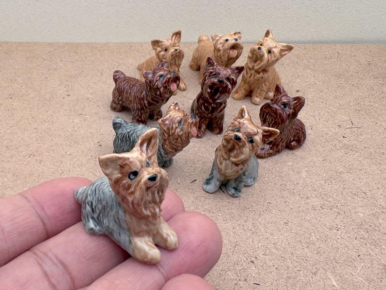 Puede incluir: Una colecci&oacute;n de nueve figuritas de cer&aacute;mica de Yorkshire Terrier en varias poses. Las figuritas est&aacute;n pintadas en tonos de marr&oacute;n, negro y blanco.