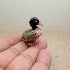 Puede incluir: Pequeña figura de pato de cerámica pintada a mano. El pato tiene cabeza negra, pico rosa y cuerpo con tonos grises, rosas y marrones. La figura está detallada con pequeños pies y se sostiene en una mano.