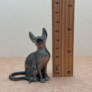 Puede incluir: Una peque&ntilde;a figurita de cer&aacute;mica de un gato sin pelo gris sentado con la cola enroscada alrededor de sus patas. El gato tiene ojos verdes y mide aproximadamente 10 cm de alto.
