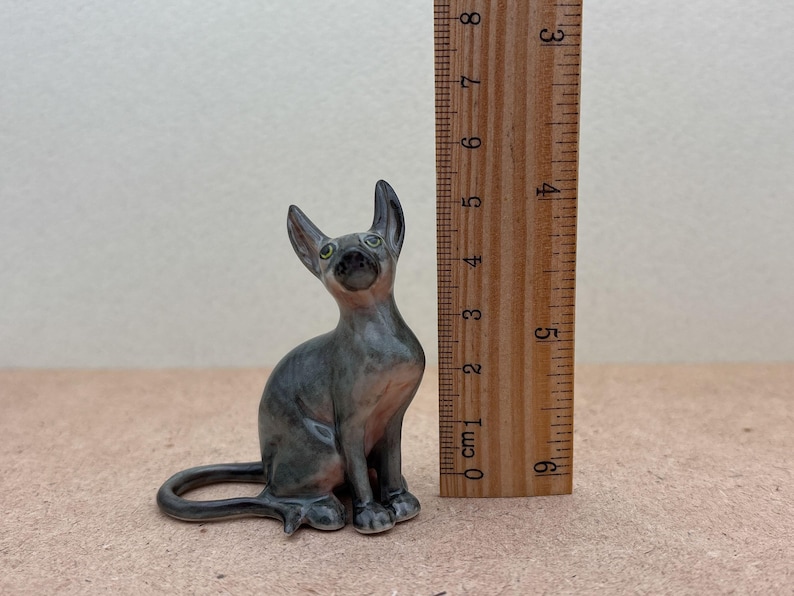 Puede incluir: Una peque&ntilde;a figurita de cer&aacute;mica de un gato sin pelo gris sentado con la cola enroscada alrededor de sus patas. El gato tiene ojos verdes y mide aproximadamente 10 cm de alto.