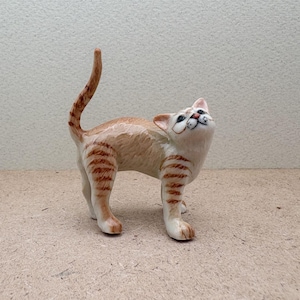 K&ouml;nnte beinhalten: Keramikfigur einer Katze mit cremefarbenem K&ouml;rper und orangefarbenen Tigerstreifen. Die Katze blickt nach oben, der Schwanz ist erhoben. Die Figur hat ein detailliertes Gesicht mit schwarzen Augen und einer rosa Nase.