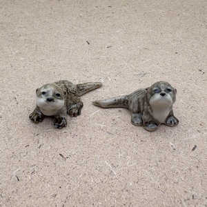 May include: Two ceramic otter figurines. The otters are in a resting position, with their bodies and tails extended. The figurines have a light brown and grey colour scheme with white faces and chests. The otters are on a light brown surface.
