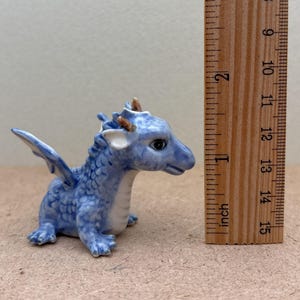 May include: A small blue ceramic dragon figurine with white accents and brown horns. The dragon is sitting with its wings spread out. The figurine is about 1 inch tall.