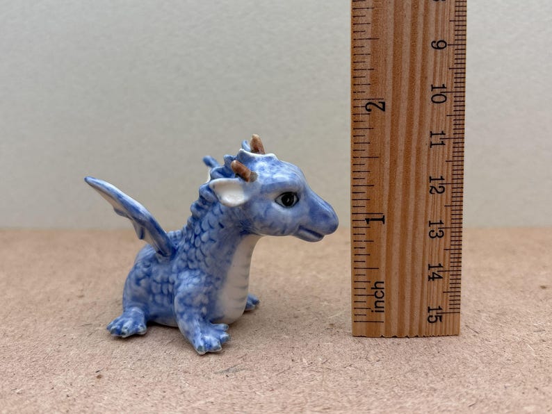 May include: A small blue ceramic dragon figurine with white accents and brown horns. The dragon is sitting with its wings spread out. The figurine is about 1 inch tall.