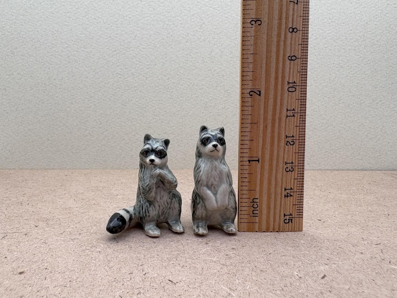 Pu&ograve; includere: Due statuine di procione in ceramica, una in piedi e una seduta, con occhi neri e un motivo a strisce grigio e bianco. Le statuine sono alte circa 5 cm e sono mostrate accanto a un righello.