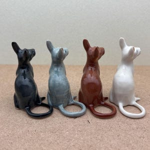K&ouml;nnte beinhalten: Vier Keramik-Katzenfiguren in verschiedenen Farben: Schwarz, Grau, Braun und Wei&szlig;. Jede Figur sitzt mit dem Schwanz um den K&ouml;rper gewickelt.