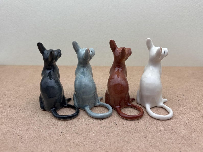 K&ouml;nnte beinhalten: Vier Keramik-Katzenfiguren in verschiedenen Farben: Schwarz, Grau, Braun und Wei&szlig;. Jede Figur sitzt mit dem Schwanz um den K&ouml;rper gewickelt.
