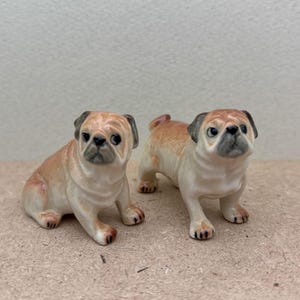 May include: Two ceramic pug dog figurines. One is sitting, the other is standing. Both have tan and cream-colored bodies with black accents on their ears and faces. The dogs have black eyes and are posed facing forward.