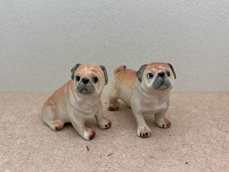 May include: Two ceramic pug dog figurines. One is sitting, the other is standing. Both have tan and cream-colored bodies with black accents on their ears and faces. The dogs have black eyes and are posed facing forward.
