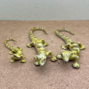 Op de afbeelding: Drie keramische gecko beeldjes in verschillende maten. De gecko's zijn geel met bruine vlekken en hebben gekrulde staarten. Ze zijn geplaatst op een lichtbruin oppervlak.