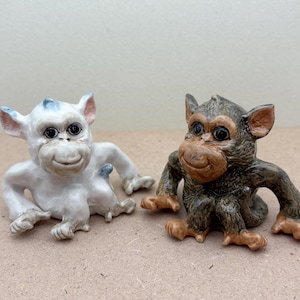 以下が含まれることがあります： 白い陶器製の猿の置物と、茶色の陶器製の猿の置物。どちらも青いアクセントと黒いアクセントが付いています。両方の猿は足を組んで座っており、友好的な表情をしています。