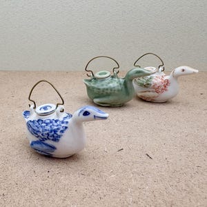May include: Three small ceramic teapots shaped like ducks. One is green with white accents, one is white with blue accents, and one is white with pink accents.